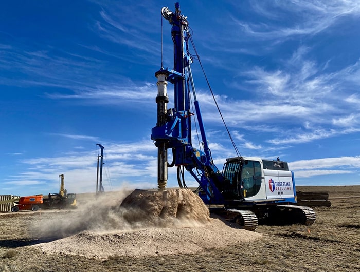A blue and white drilling rig operating on a pile of dirt at a construction site.