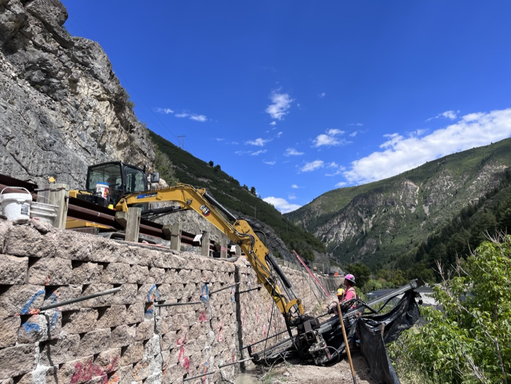 Construction workers using drilling equipment on a rock wall for a short-term project. Rental equipment is being utilized.
