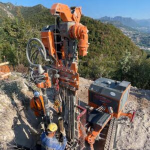 A man standing next to an orange drilling rig on a hillside.