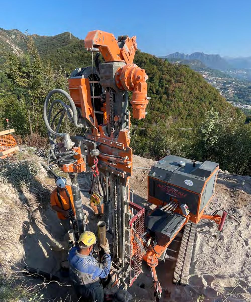 A man standing next to an orange drilling rig on a hillside.