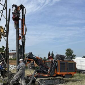 A man in orange coveralls works on a drill rig in front of a power pole.