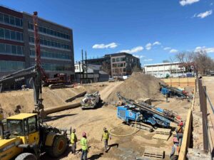 Construction site with workers, heavy machinery, and a large pile of dirt near modern buildings.