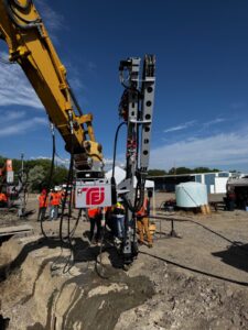 Construction workers operating a drilling rig with an excavator in an outdoor site.