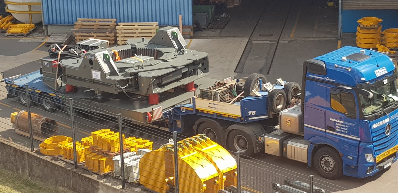 Large industrial machinery being transported on a blue flatbed truck in an industrial yard with yellow equipment parts nearby.