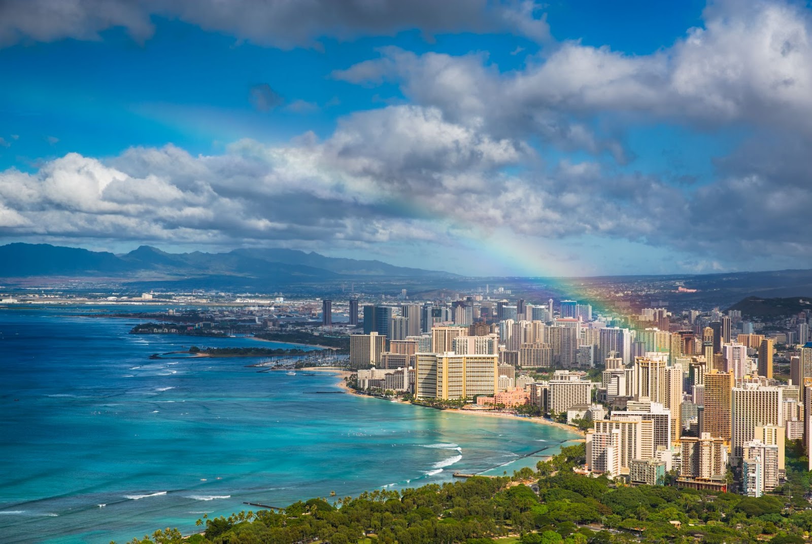 Scenic view of Honolulu’s coastline with turquoise waters, high-rise buildings, and a rainbow stretching across the city.