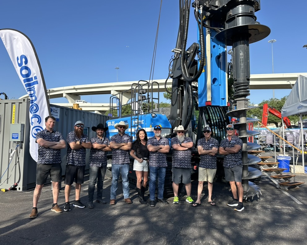 Group of 9 people posing in front of a large blue Soilmec SR-30 drilling rig under a highway overpass. Most wear matching patterned shirts; one woman in black top. Sunny day, construction site vibe