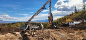 Construction site with CALCO concrete pump truck pouring foundation piers, drill rig boring holes, and pickup trucks parked on a scenic mountain hillside with autumn trees and clear blue sky.