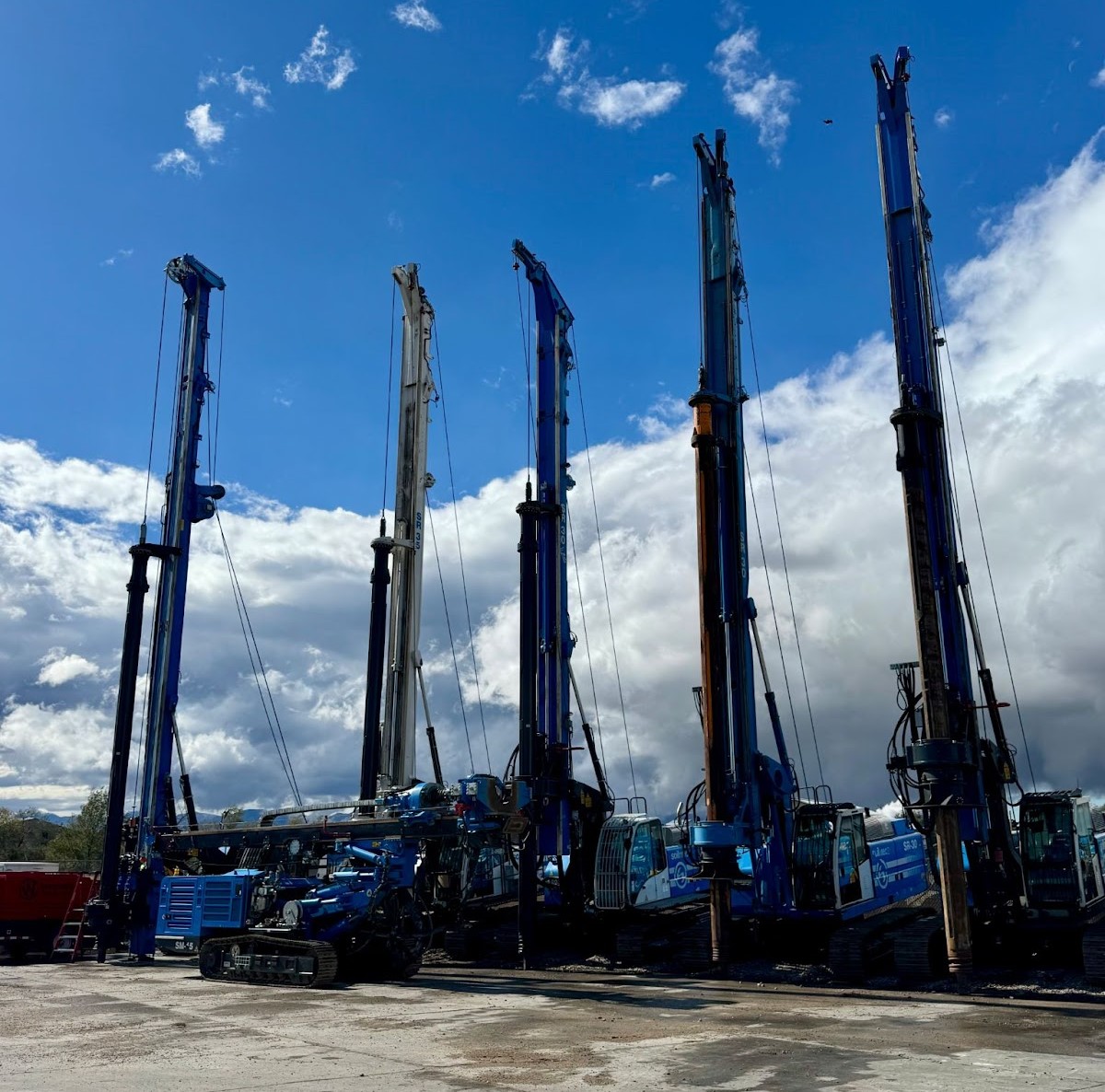 Five blue pile driving rigs parked in a row on a concrete lot under a bright blue sky with scattered clouds. Heavy construction machinery ready for foundation work.