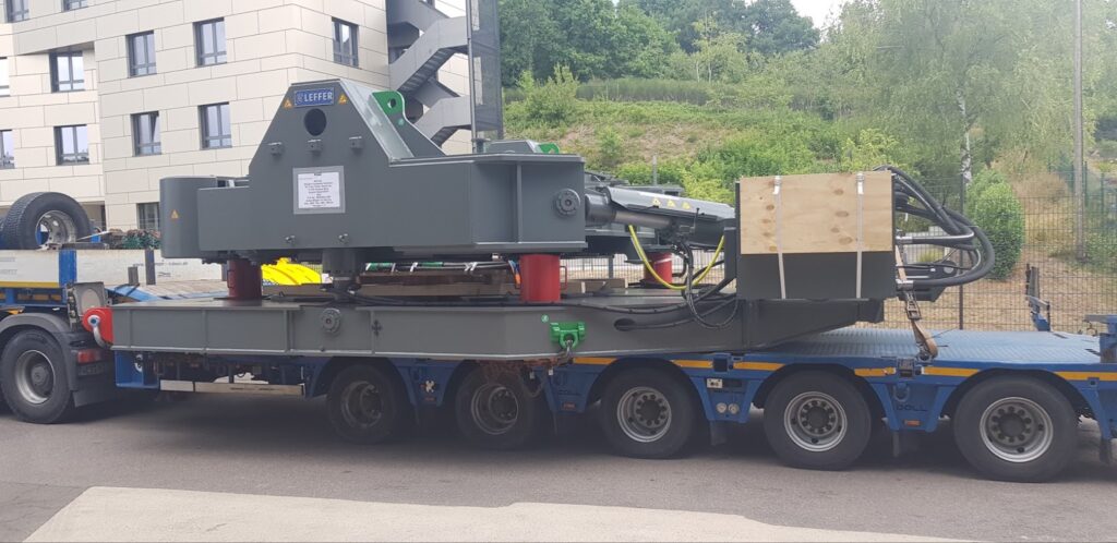 Heavy machinery, possibly a crane or industrial equipment, being transported on a large flatbed truck near a building and greenery.
