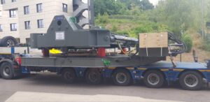 Heavy machinery, possibly a crane or industrial equipment, being transported on a large flatbed truck near a building and greenery.