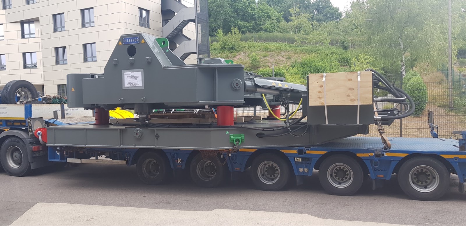 Heavy machinery, possibly a crane or industrial equipment, being transported on a large flatbed truck near a building and greenery.