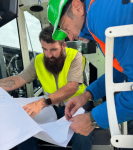 Two workers in safety gear reviewing blueprints inside a vehicle.