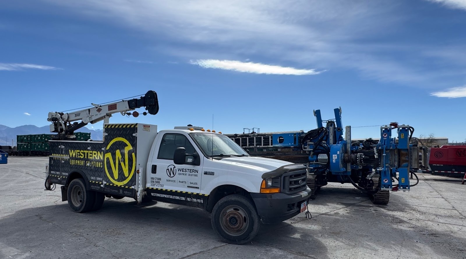 White truck with Western Equipment Solutions branding and a crane, parked next to blue machinery under a clear sky.