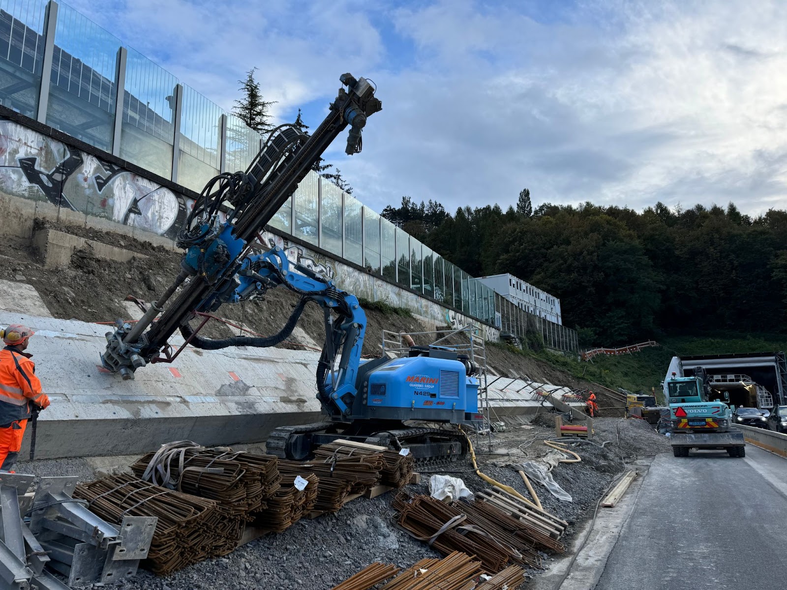 A worker in an orange safety suit operates a blue Marini Quarries Group crawler drilling rig (model N429) as it bores into a sloped embankment for ground anchoring or soil nailing during roadside slope stabilization on a highway construction site.