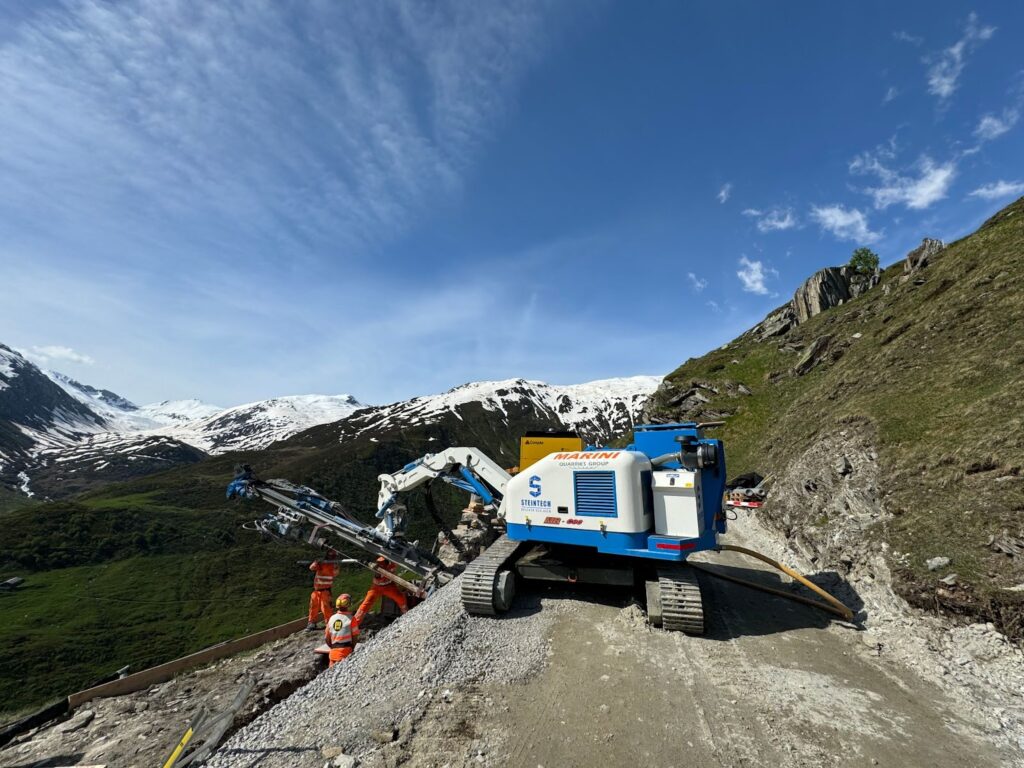 Workers in high-visibility orange suits operate a tracked drilling rig from Marini Quarries Group (labeled ZANETTI QUARRIES GROUP and MARINI 600) at a high-altitude site in the Swiss Alps.