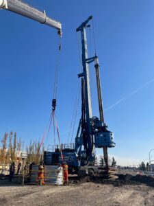 Construction workers using a crane and a large blue drilling rig to install a corrugated metal culvert at a construction site.