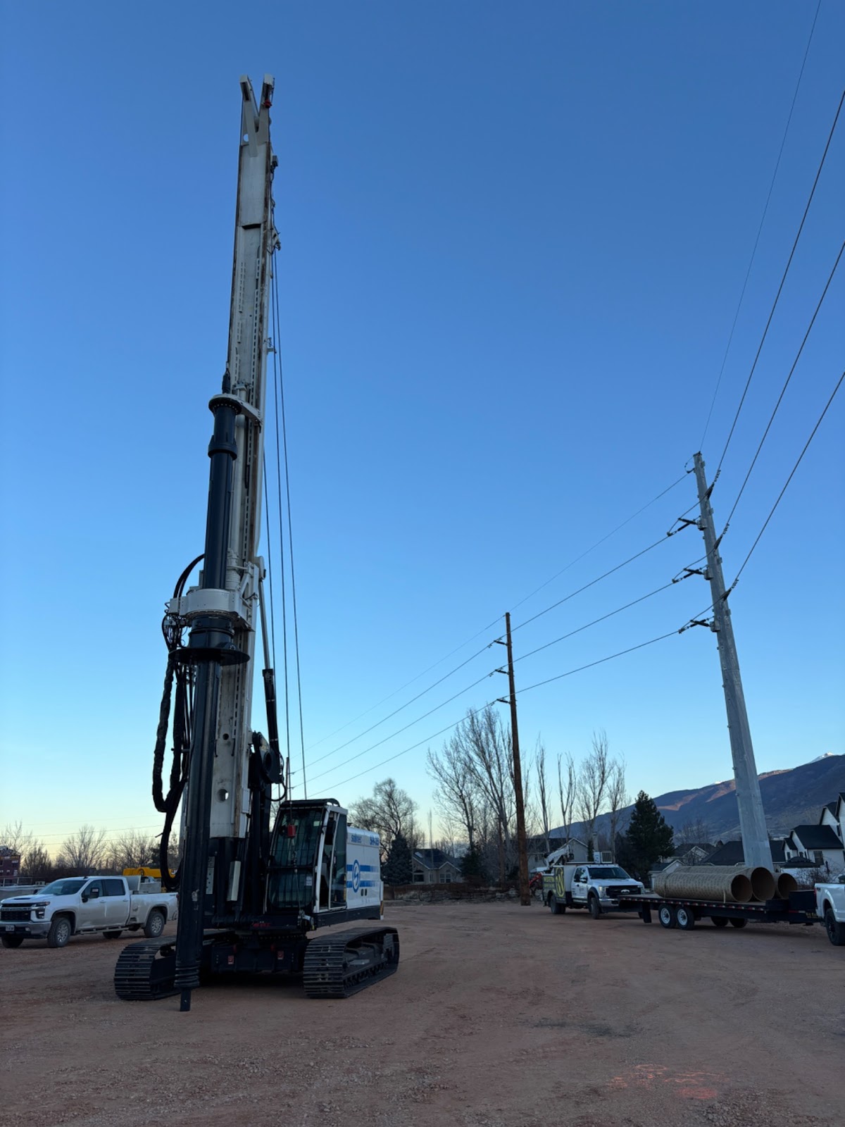 A Soilmec drilling rig parked at a construction site near power lines and a trailer carrying large concrete pipes.