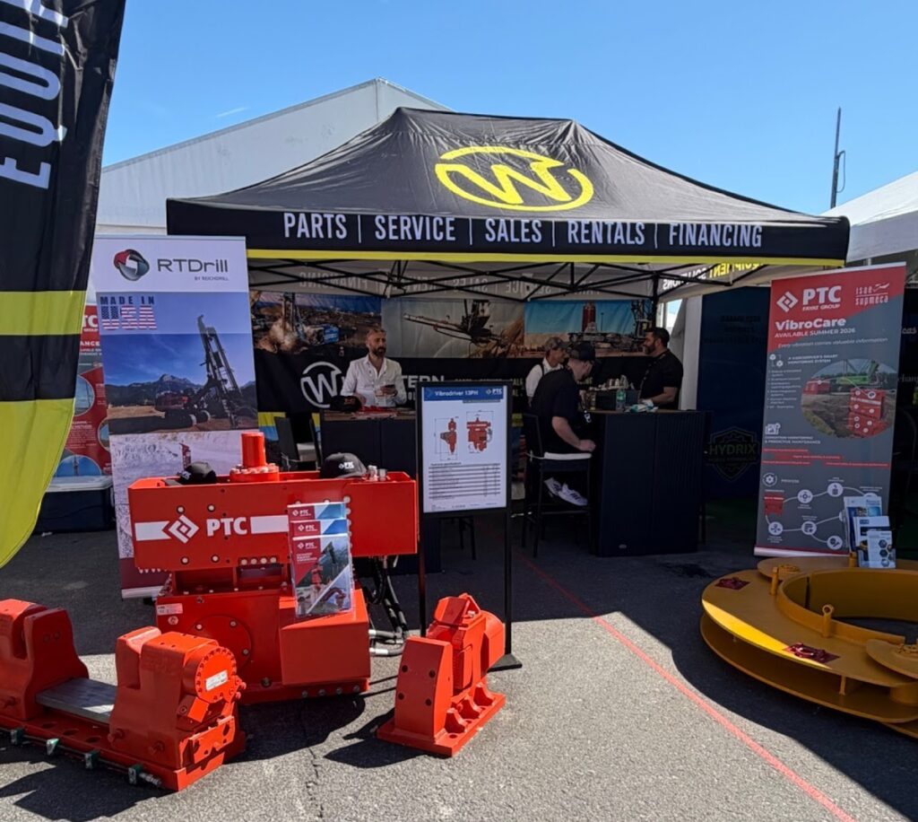 An outdoor trade show booth for Western Equipment and PTC, featuring heavy drilling machinery components, marketing banners, and staff under a black canopy tent