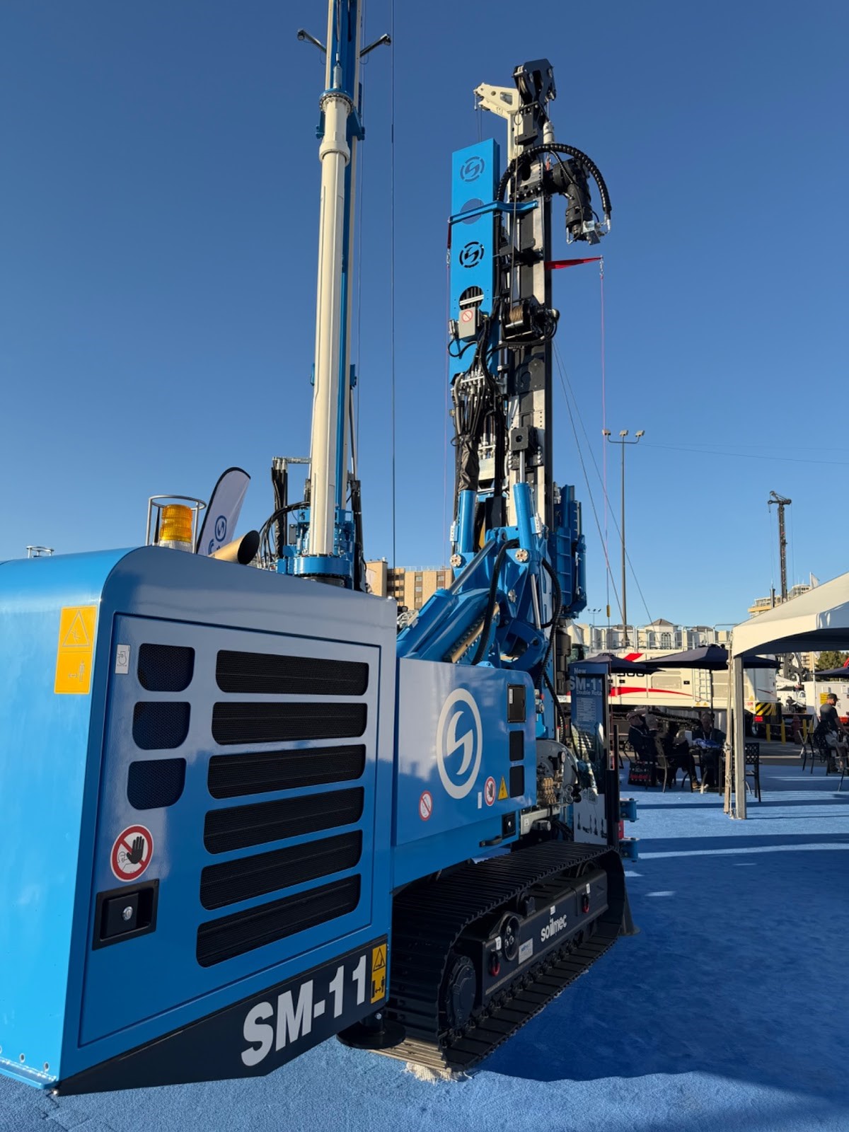 A side-view of a blue Soilmec SM-11 hydraulic drilling rig displayed on blue carpeting at an outdoor industrial trade show.