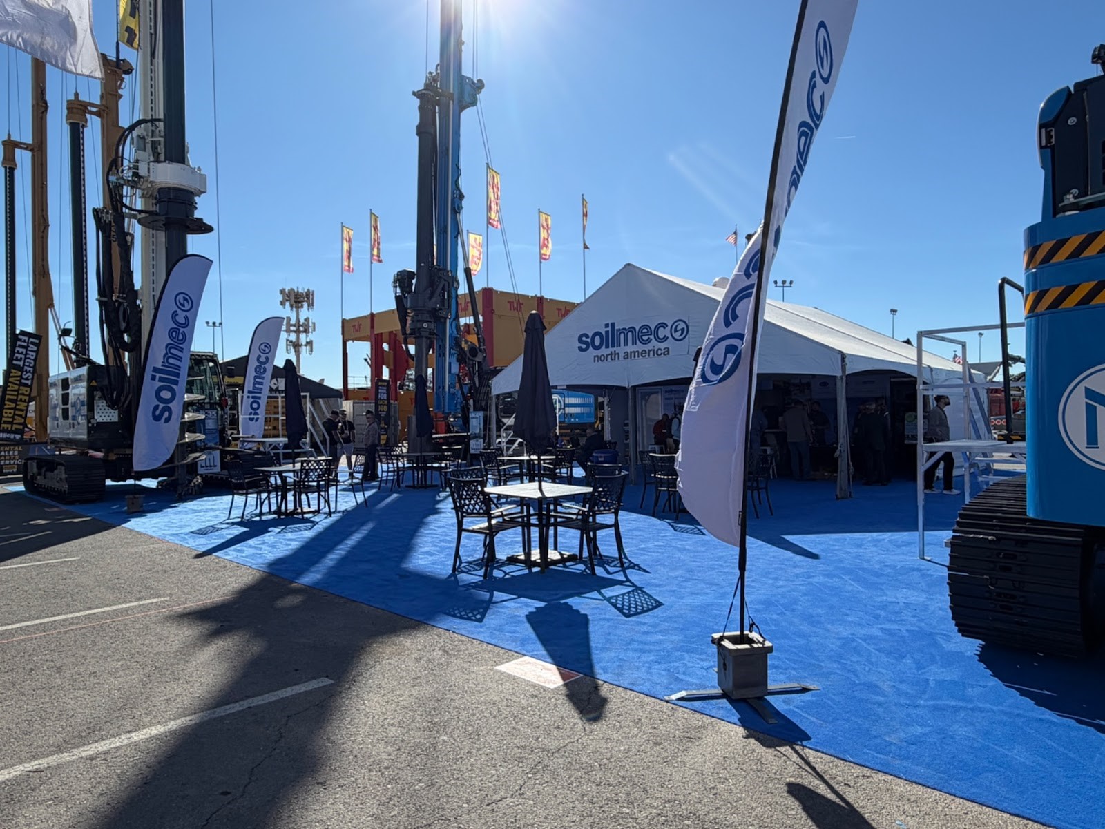 An outdoor trade show exhibit for Soilmec North America featuring heavy foundation drilling rigs, promotional flags, and a large white hospitality tent on blue carpeting.