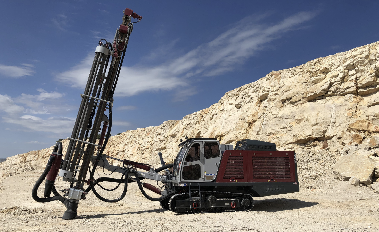A large, tracked drilling rig operating in an open-pit quarry against a blue sky.