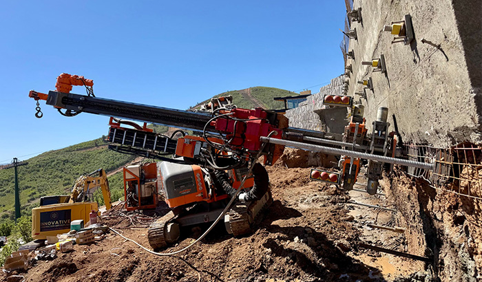 A large tracked drilling rig operating on a dirt slope to install tiebacks or anchors into a concrete retaining wall.