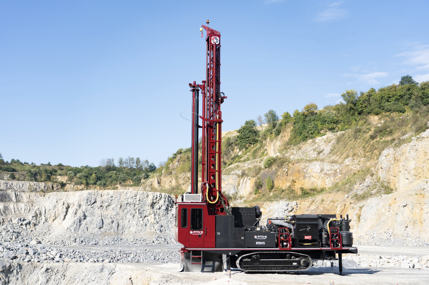 A red and black tracked blast hole drill rig positioned on a rocky bench inside an open-pit quarry.