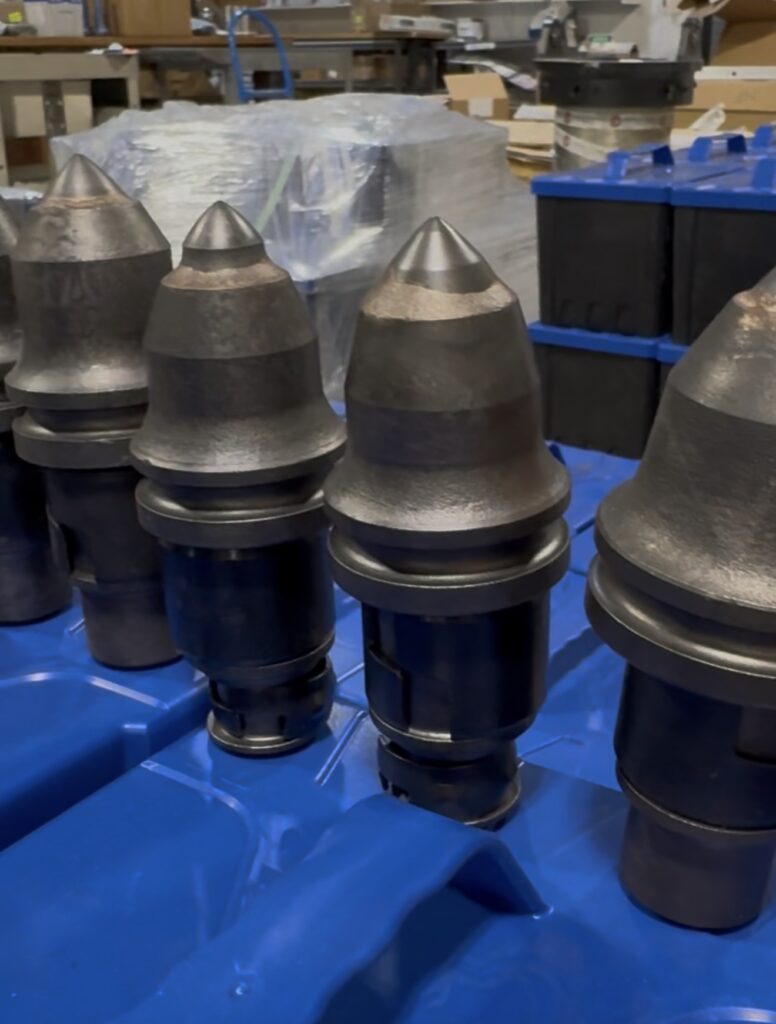 A row of heavy-duty, conical metal drill bits or cutting teeth sitting upright on a bright blue surface.