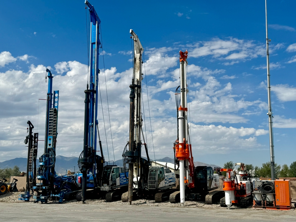 A row of several large, industrial foundation drilling rigs parked outdoors under a blue, partly cloudy sky.