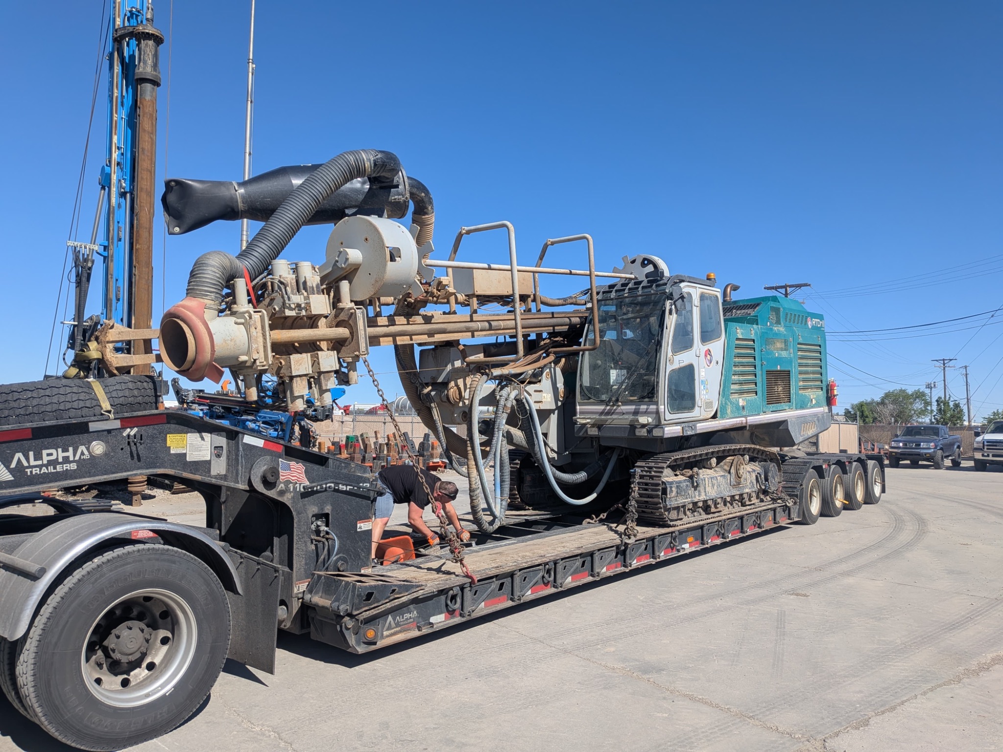 A worker secures a large, teal-and-white tracked drilling rig onto a lowboy flatbed trailer.