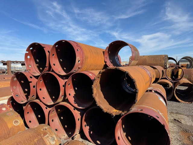 A stack of large, rusty industrial pipes and core barrels with circular holes around the rims, sitting outdoors under a blue sky with light clouds.