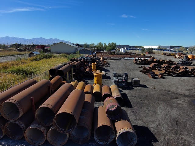 An outdoor industrial yard filled with scattered stacks of large, rusty metal casings, pipes, and drilling equipment under a clear blue sky.
