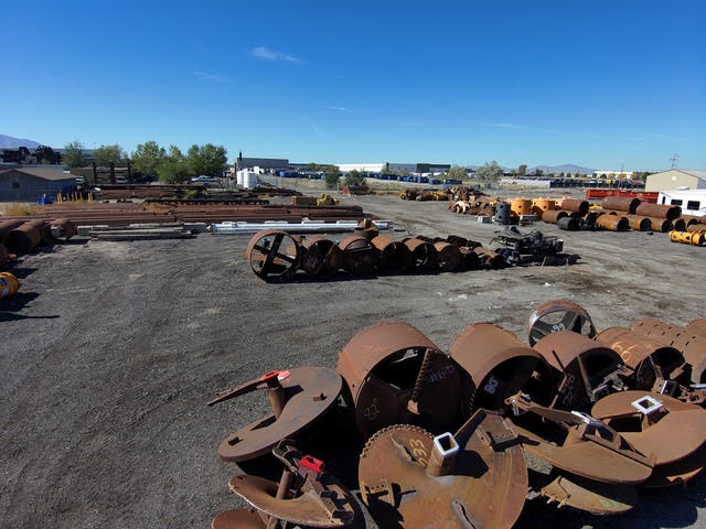 An outdoor storage yard filled with various heavy industrial drilling equipment, including rusty augers, core barrels, and large metal casings, under a clear blue sky.