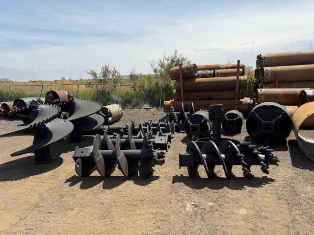 Black industrial drilling augers and flighting resting on the ground in an outdoor yard, with stacked rusty pipes in the background.