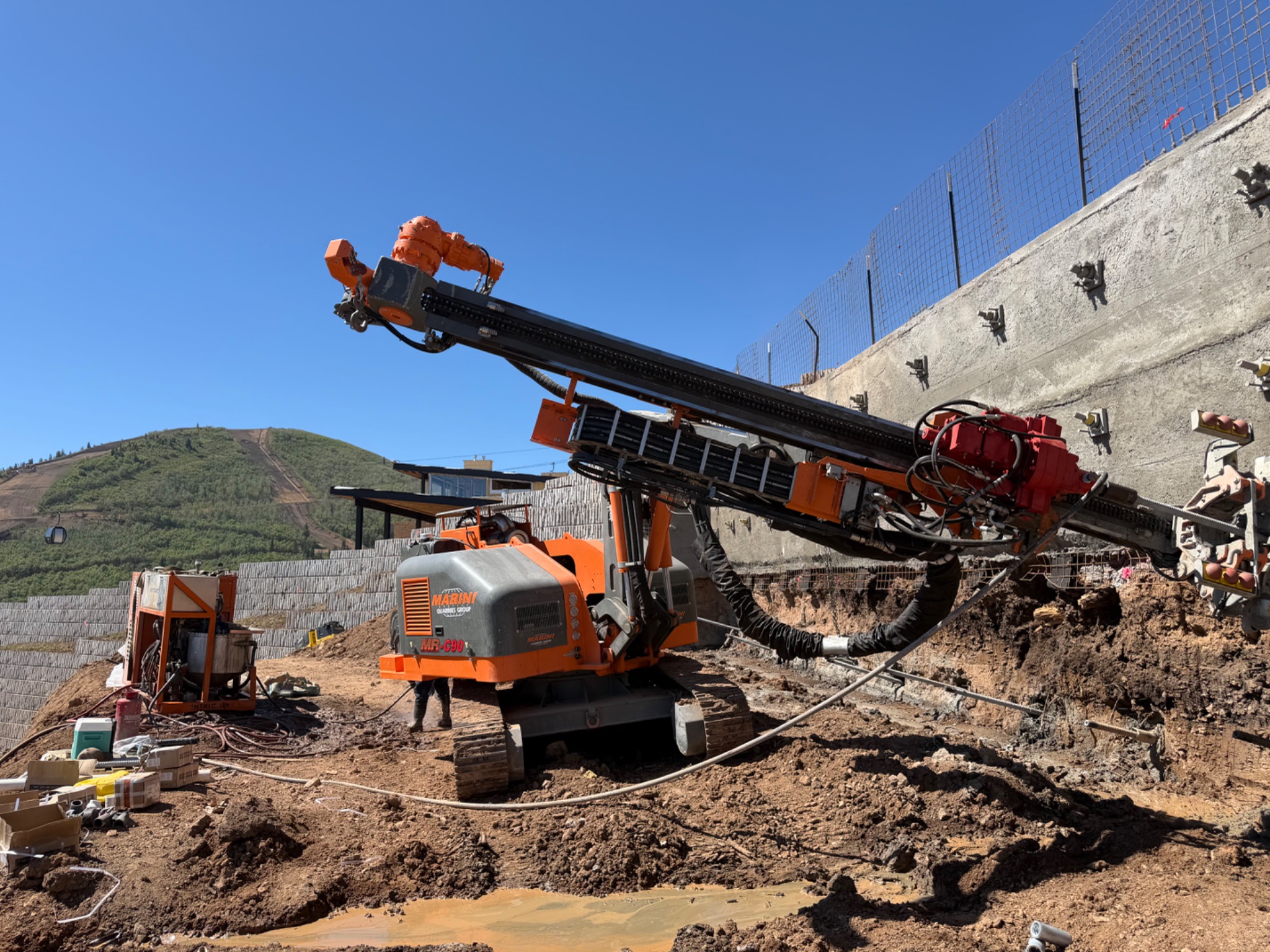 Orange tracked drilling rig machine performing tieback or anchor drilling along a reinforced slope at a mountain construction site.