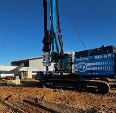 Soilmec SR-65 drilling rig at a construction site near an American Airlines airplane, with foundation drilling rigs in operation and industrial equipment in the background.